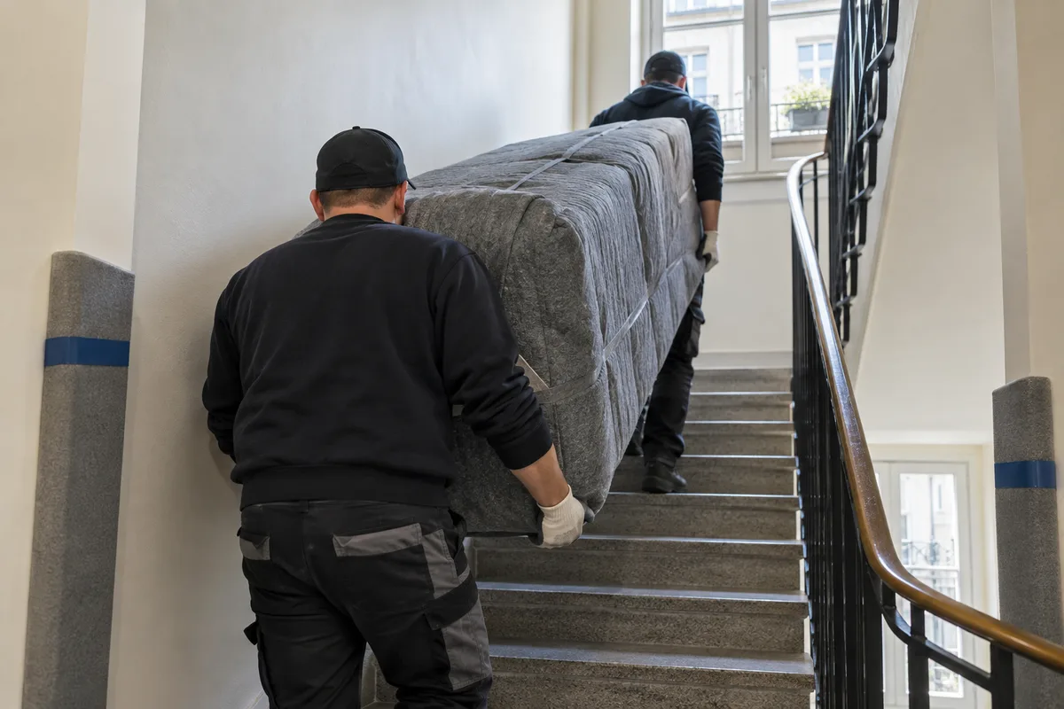 Deux déménageurs vus de dos portant un canapé emballé dans une cage d’escalier claire